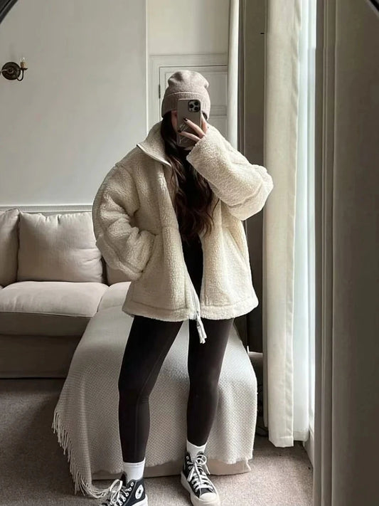 Person wearing a beige coat, black leggings, and black sneakers in a room with a couch and white walls.