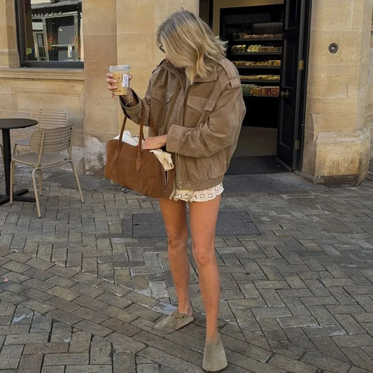 Woman in a brown jacket and shorts holding a drink on a paved street.