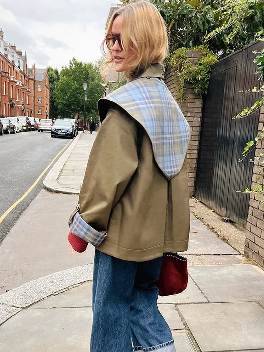 Woman walking on a street wearing a green jacket with plaid lining, blue jeans, and a red bag.