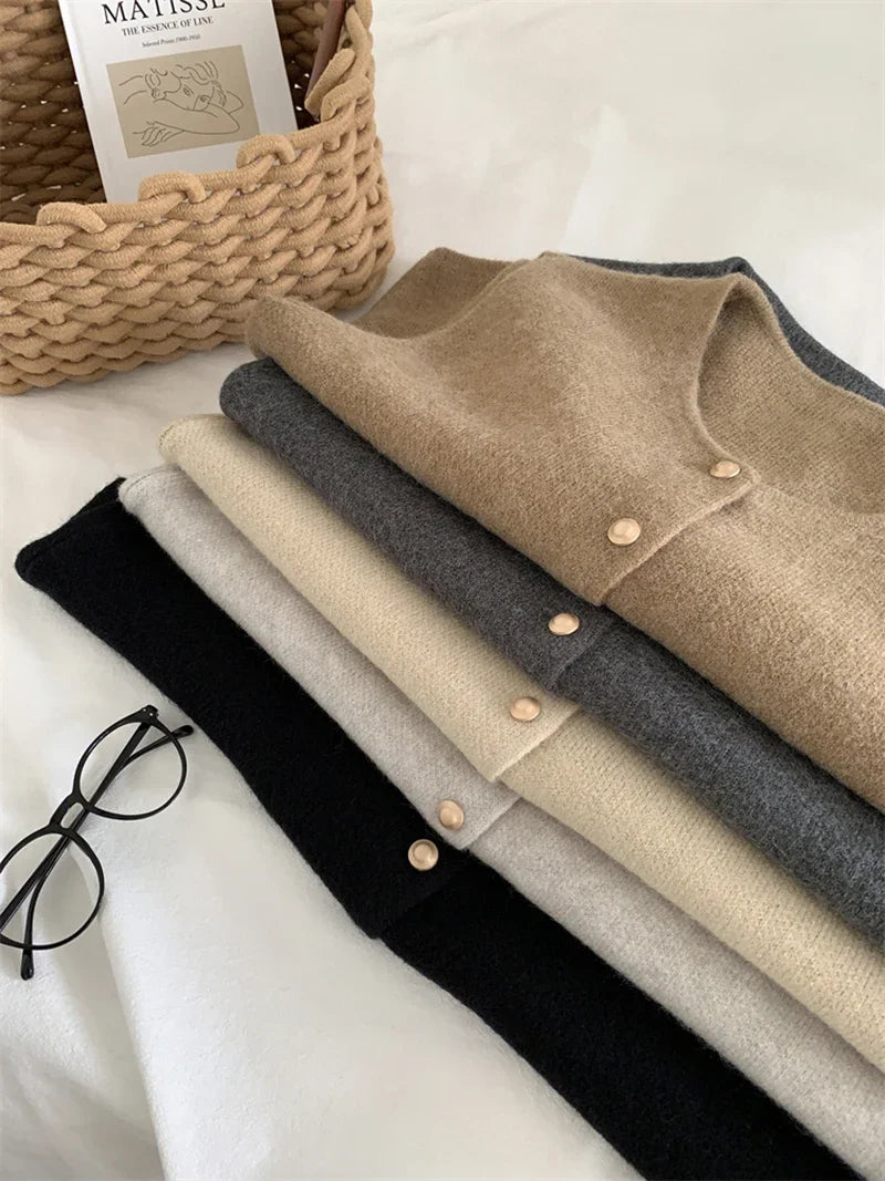 Stack of folded sweaters in black, gray, and beige on a white surface with a basket and book in the background.