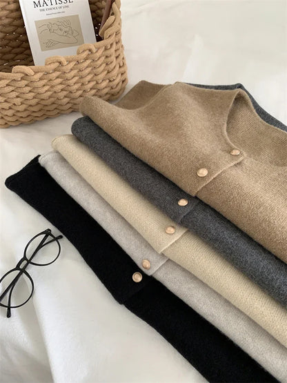 Stack of folded sweaters in black, gray, and beige on a white surface with a basket and book in the background.