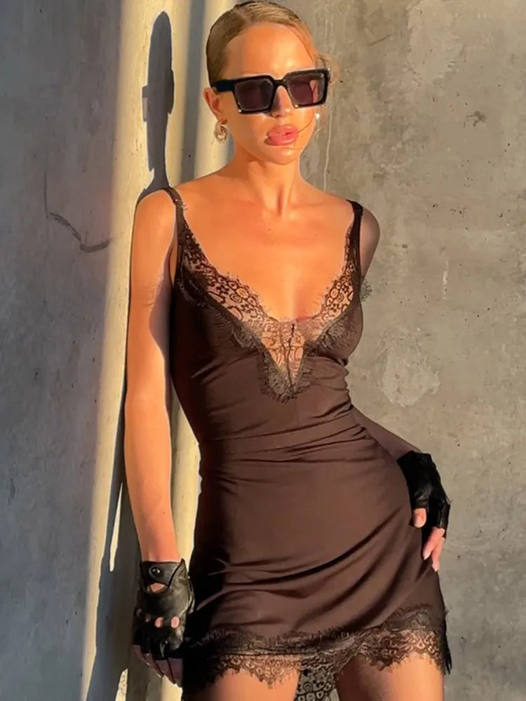 Woman wearing a brown lace dress and sunglasses against a gray wall.