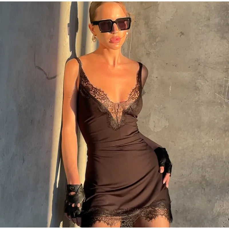 Woman wearing a brown lace dress and sunglasses against a gray wall.