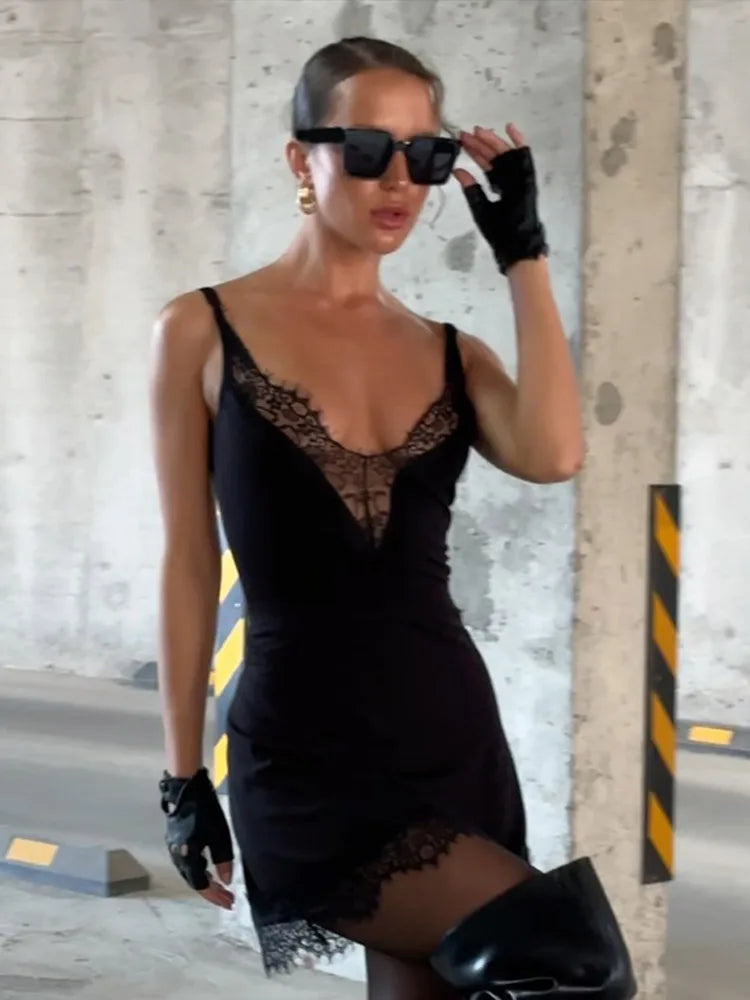 Woman in a black dress with lace details, sunglasses, and gloves standing against a concrete wall.