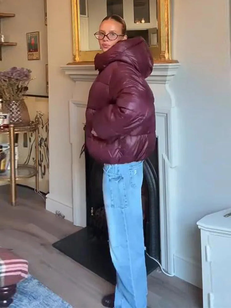Person wearing a maroon puffer jacket and blue jeans standing in a room with a fireplace.
