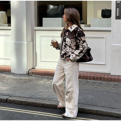 Woman in a patterned shirt and white pants standing on a street, holding a drink.