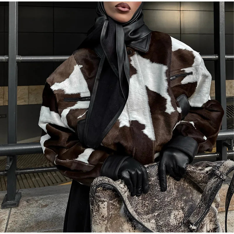 Person wearing a cow print jacket with black gloves and a scarf, sitting on a bench.
