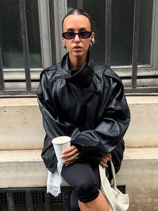 Person wearing a black leather jacket and sunglasses, sitting on steps holding a coffee cup.