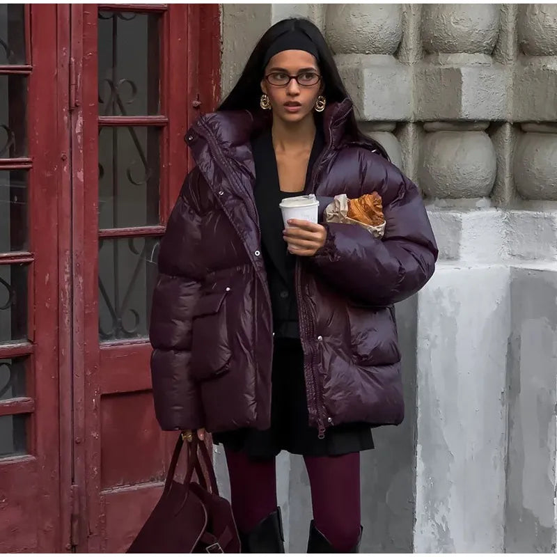 Person wearing a purple puffer jacket holding a coffee cup and pastry against a textured wall.