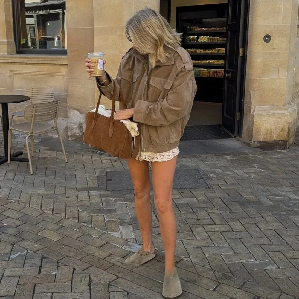 Woman in a brown jacket and shorts holding a drink on a paved street.