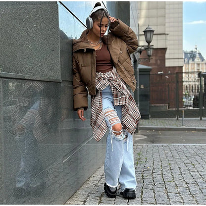 Person wearing a brown puffer jacket and ripped jeans leaning against a wall.