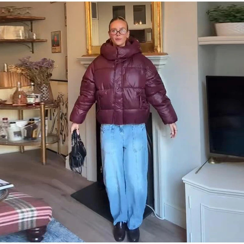Person wearing a maroon puffer jacket and blue jeans standing in a living room.