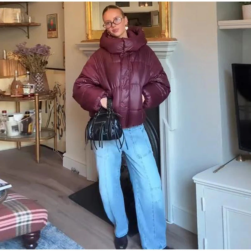 Person wearing a maroon puffer jacket and blue jeans standing in a living room.
