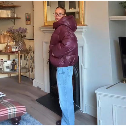 Person wearing a maroon puffer jacket and blue jeans standing in a room with a fireplace and shelves.
