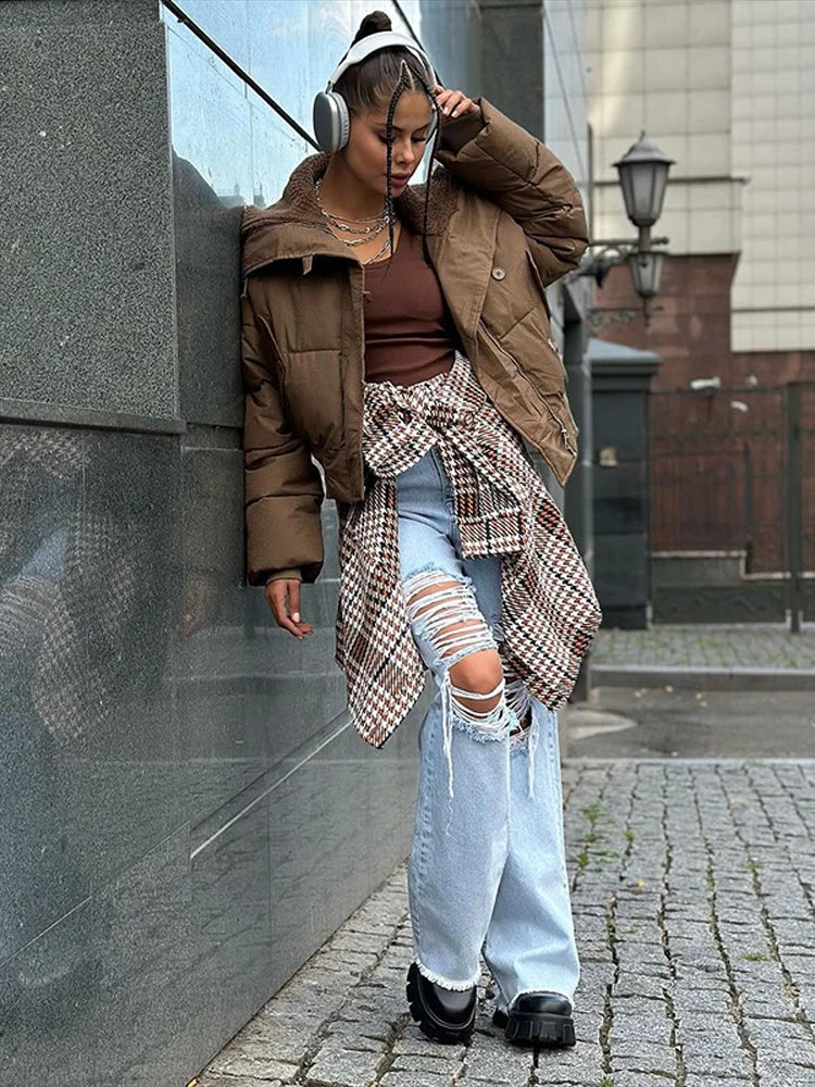 Person wearing a brown puffer jacket and ripped jeans standing against a wall.