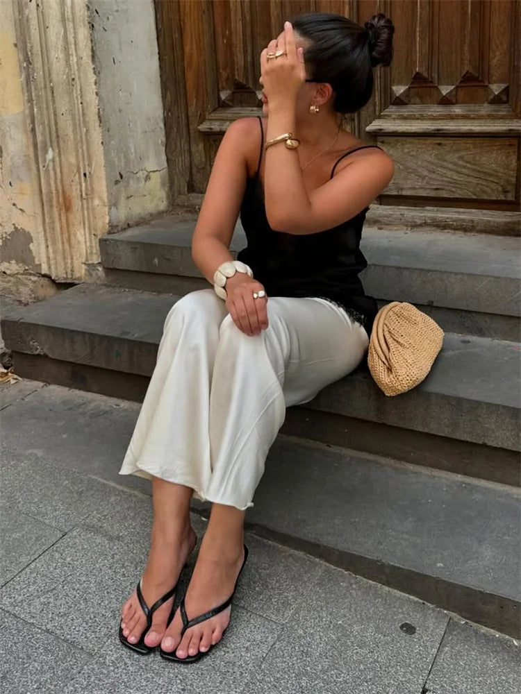 Woman sitting on stone steps wearing a black top and white pants with a straw bag beside her.