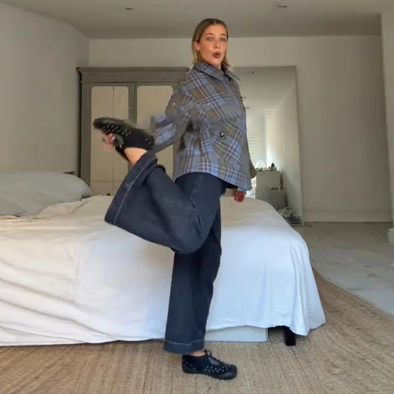 Person in plaid jacket and jeans standing in a bedroom with a bed and dresser in the background.