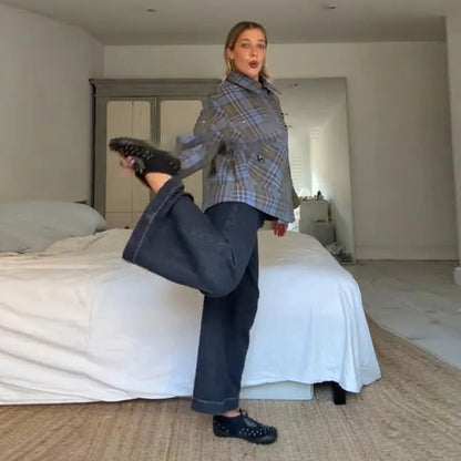 Person in plaid jacket and jeans standing in a bedroom with a bed and dresser in the background.