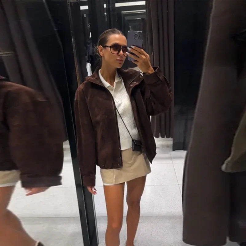 Woman taking a mirror selfie wearing a brown jacket and beige shorts.