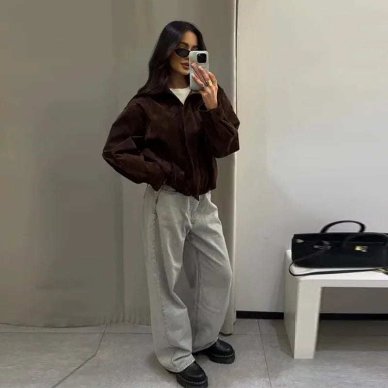 Person taking a mirror selfie wearing a brown jacket and light-colored pants in a minimalistic room.