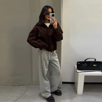 Person taking a mirror selfie wearing a brown jacket and light-colored pants in a minimalistic room.