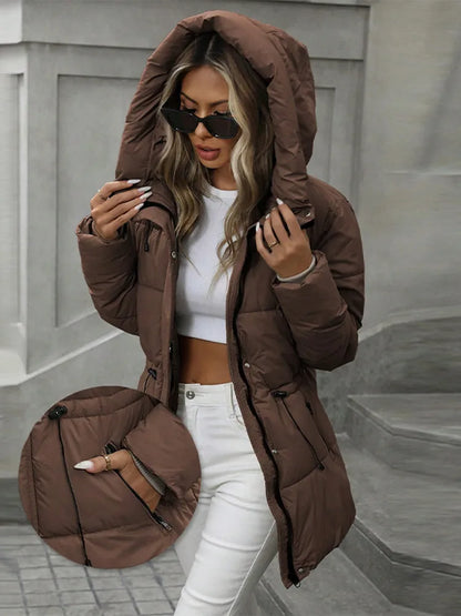 Woman wearing a brown puffer jacket with a hood, white top, and white pants on a neutral background.