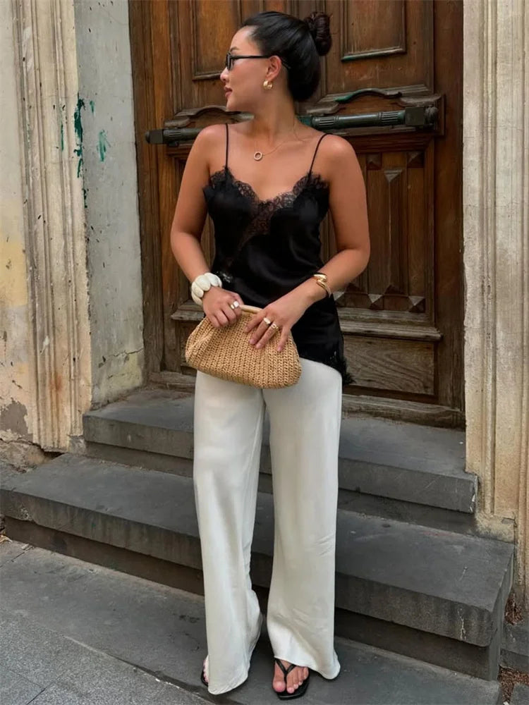 Woman in black lace top and white pants standing on steps holding a woven handbag.