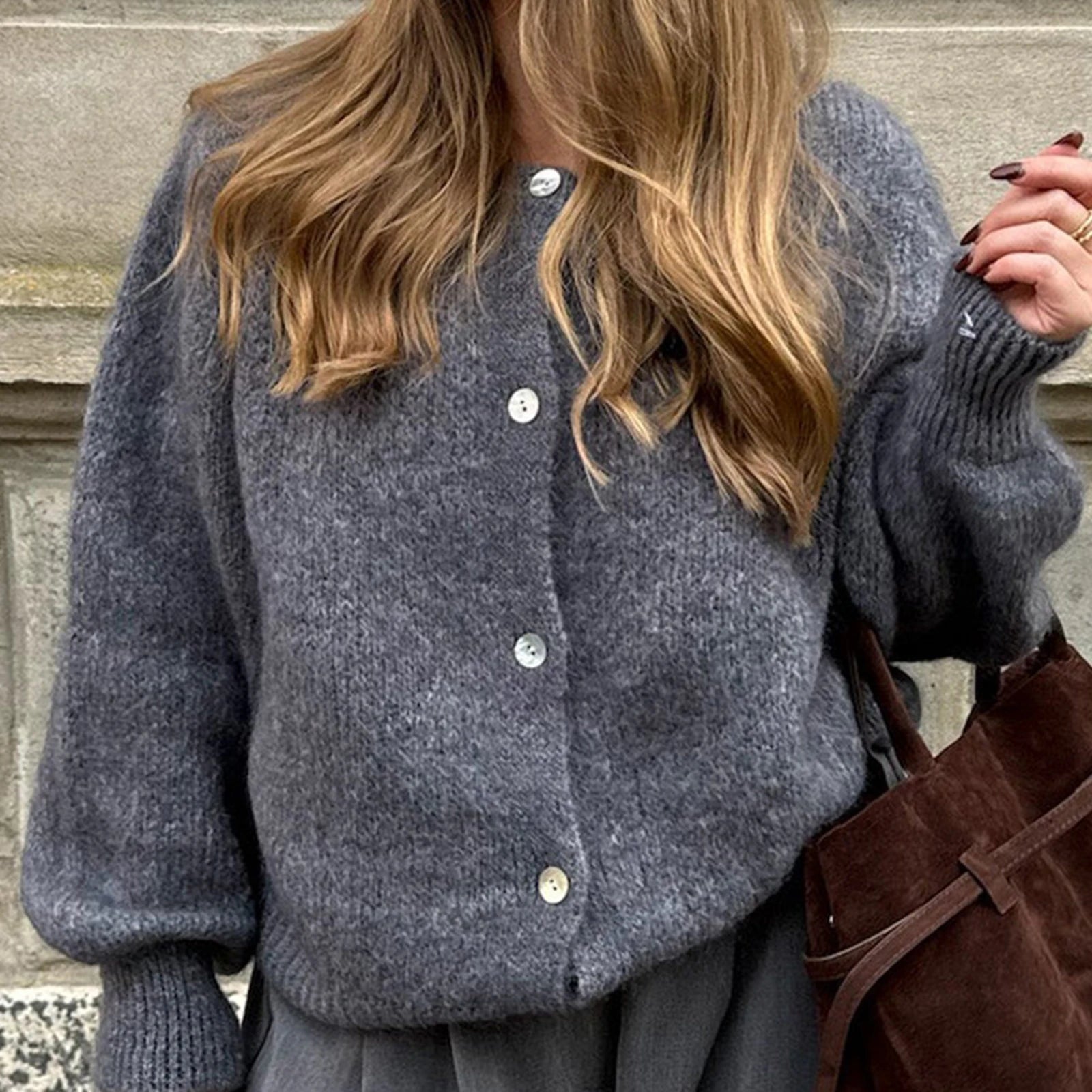 Person wearing a gray knitted cardigan with a brown bag against a stone wall.