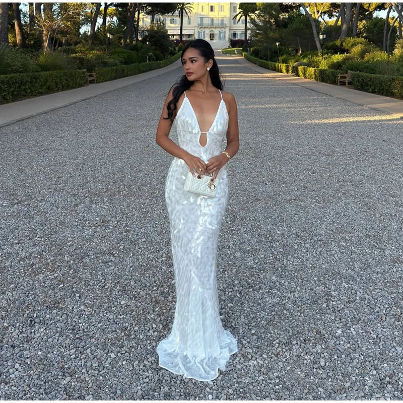 Woman in a white evening gown standing on a gravel path with greenery and a building in the background.