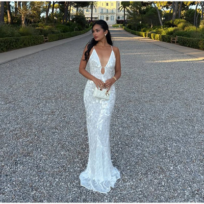 Woman in a white evening gown standing on a gravel path with greenery and a building in the background.