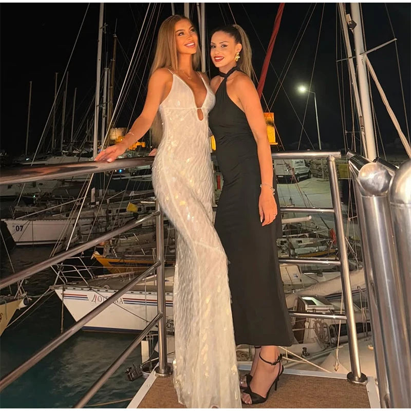 Two women in formal dresses standing on a dock with boats in the background.