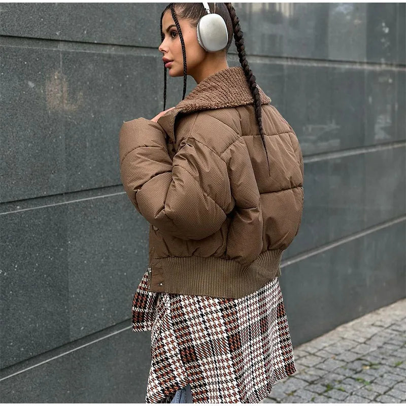 Woman wearing a brown puffer jacket and plaid skirt with headphones, standing against a gray wall.