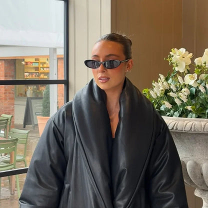 Person wearing a dark robe and sunglasses indoors with a window and plants in the background