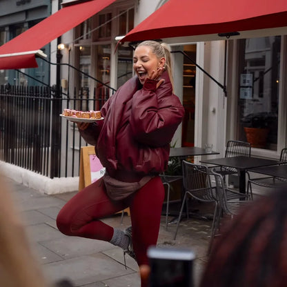 Woman in maroon jacket and leggings holding a tray of food outdoors with red awnings.