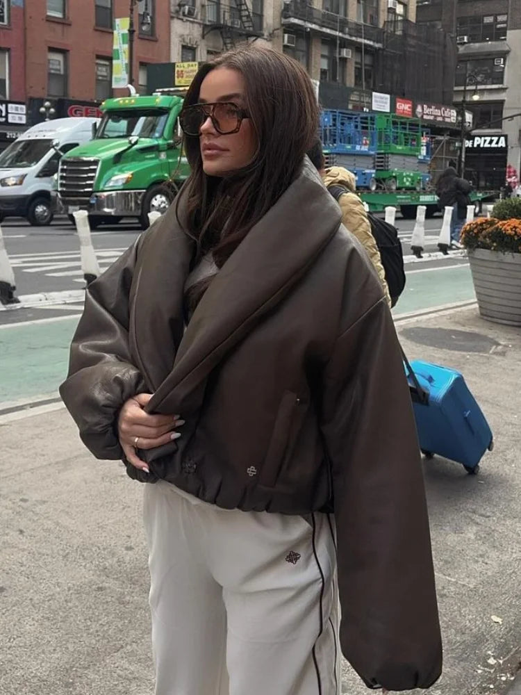 Woman in a brown coat standing on a city street with vehicles and pedestrians in the background.