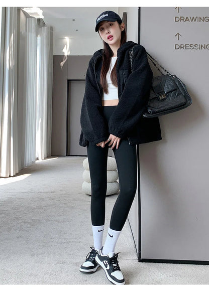 Woman in black jacket, leggings, and sneakers standing in a modern interior setting.