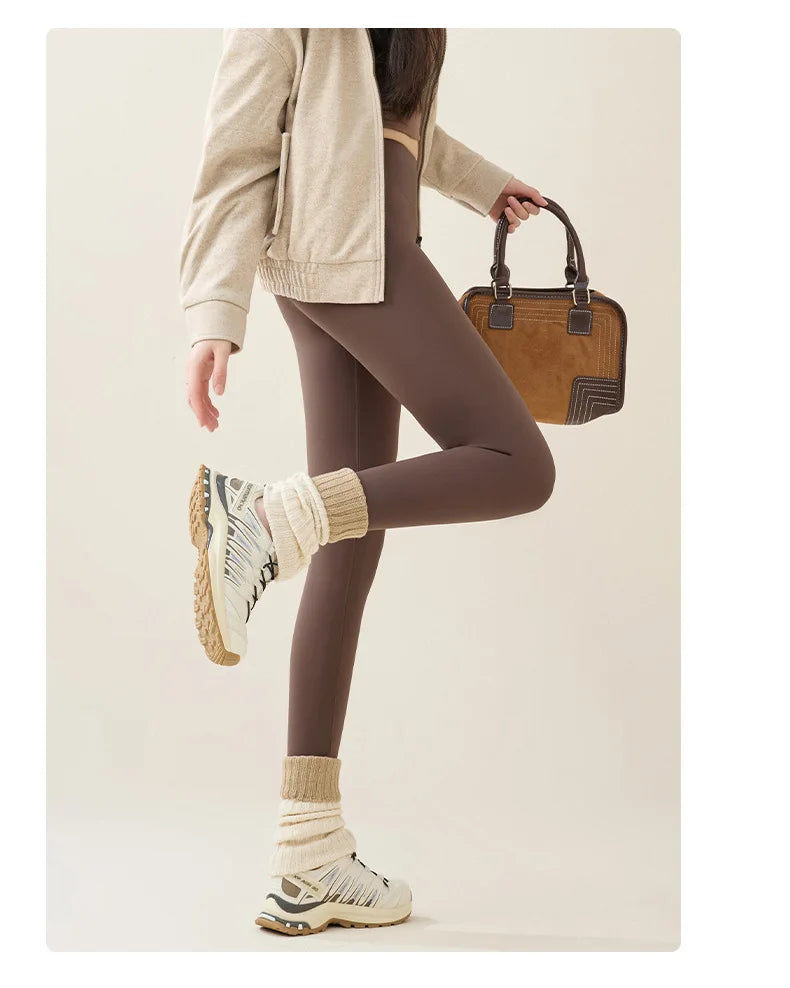 Person wearing brown leggings, beige jacket, and white sneakers holding a brown handbag on a plain background