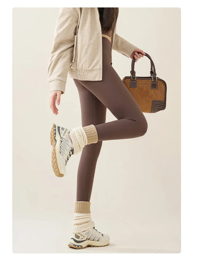 Person wearing brown leggings, beige jacket, and white sneakers holding a brown handbag on a plain background