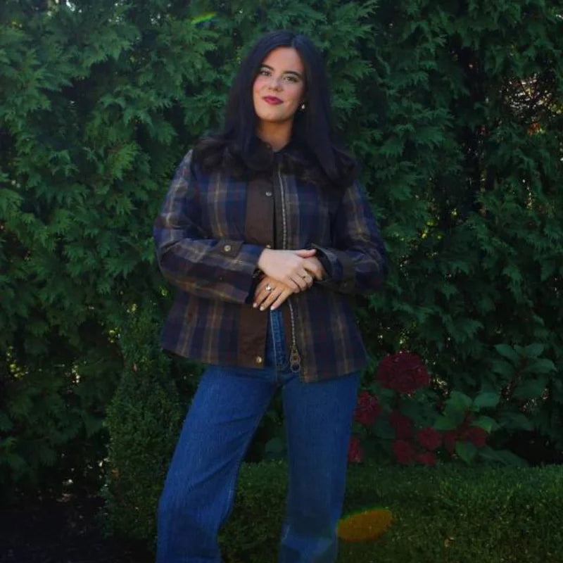 Woman wearing a plaid jacket and blue jeans standing in front of green bushes