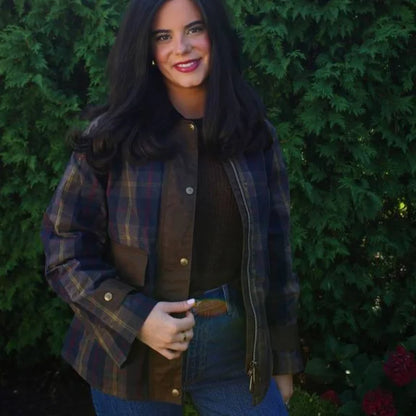 Woman wearing a plaid jacket over a brown shirt, standing against a green bush.