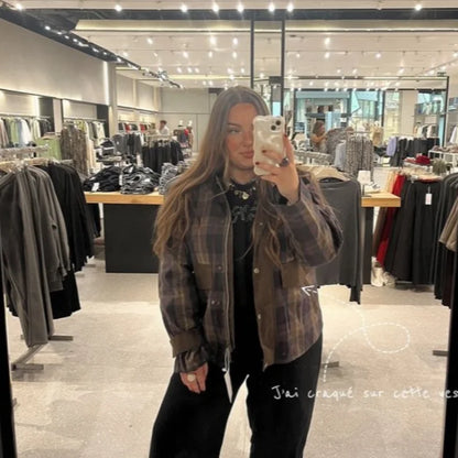 Person taking a mirror selfie in a clothing store wearing a plaid shirt and black pants.