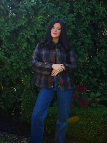 Person wearing a plaid jacket and blue jeans standing in front of green bushes.