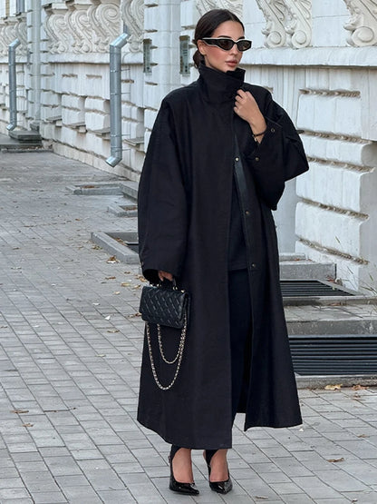 Woman in a long black coat standing on a sidewalk with decorative white building in the background