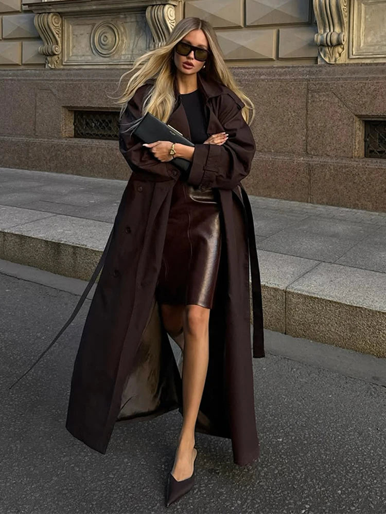 Woman in a long brown coat and leather skirt standing on a city street.