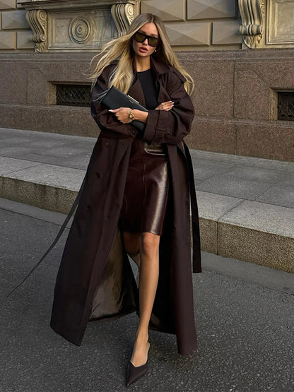Woman in a long brown coat and leather skirt standing on a city street.