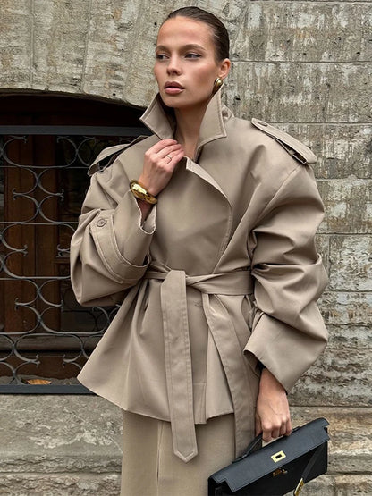Woman wearing a beige trench coat holding a black handbag against a stone wall.