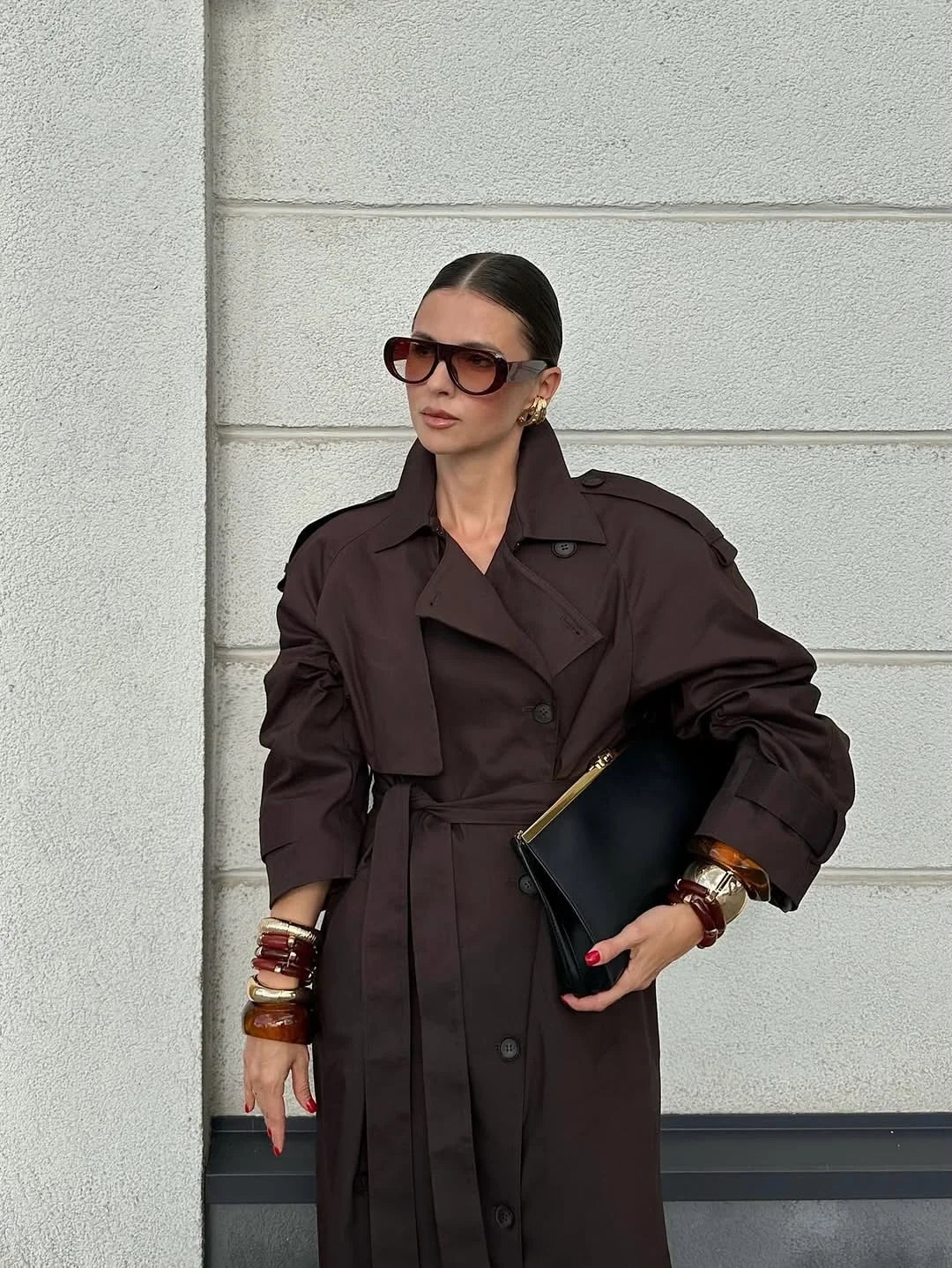 Woman wearing a brown trench coat and sunglasses against a gray wall.