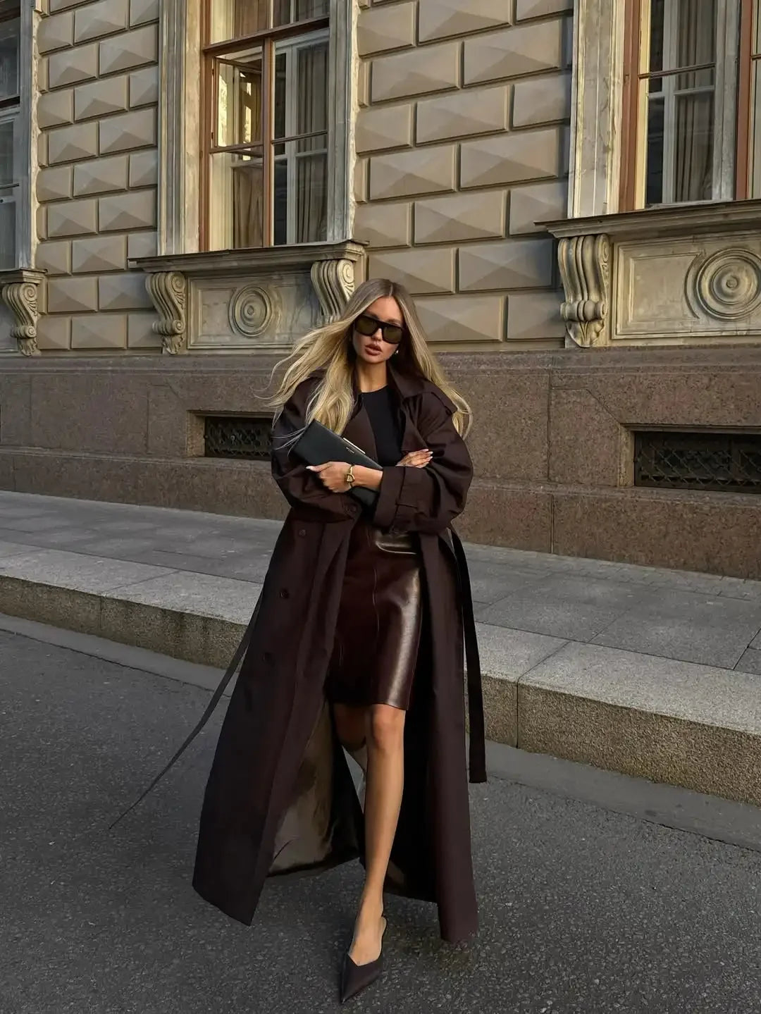 Woman in a long brown coat and black leather skirt standing in front of a building.