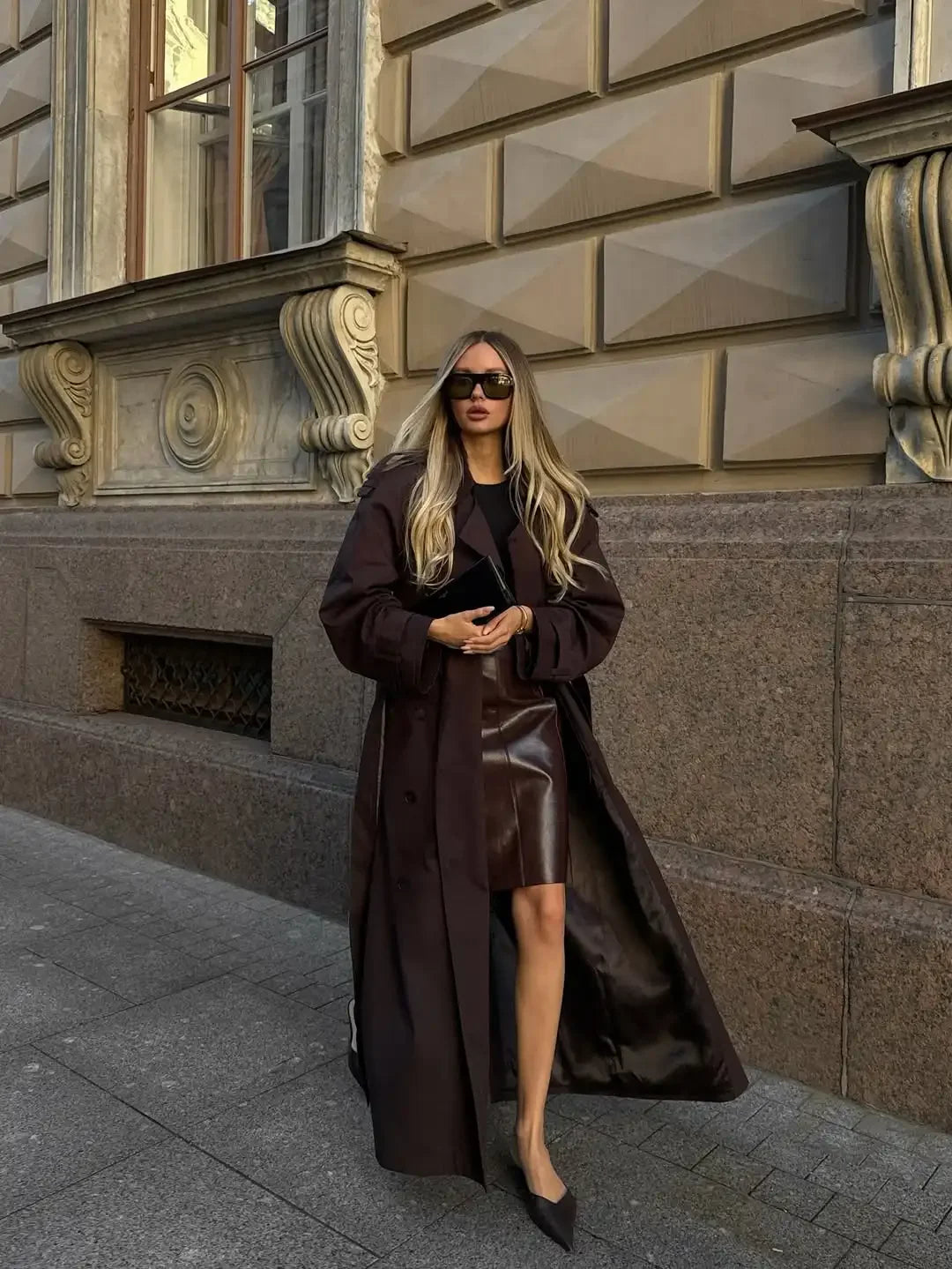 Woman in a long brown coat standing on a street with classical architecture in the background