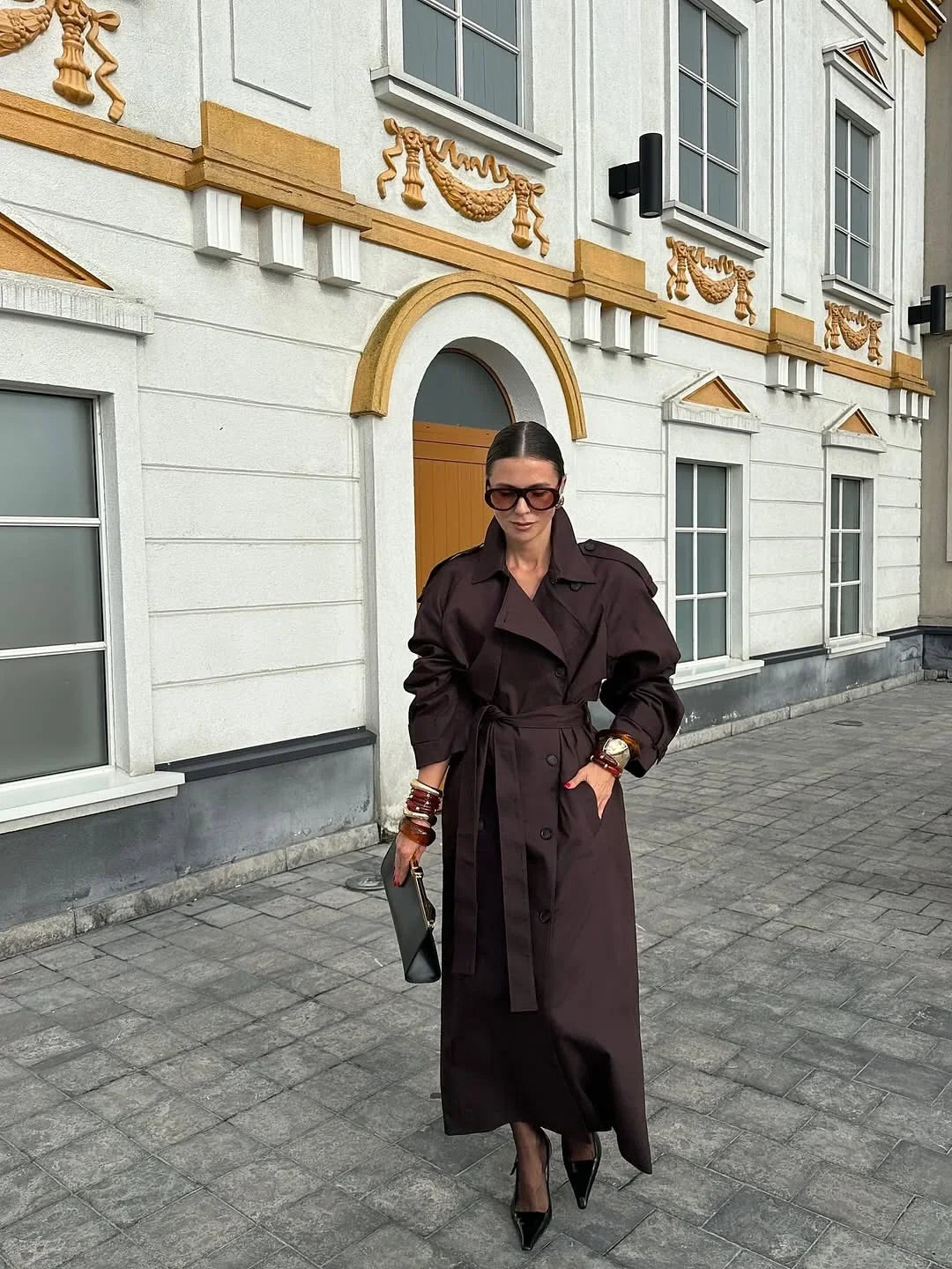 Woman in a long brown coat standing in front of an ornate building.